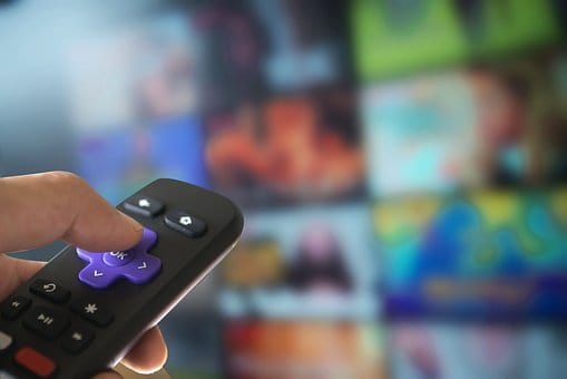 Image of a person's hand, holding a Roku remote in front of blurred screen with streaming choices.