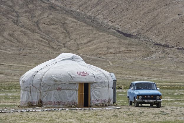 A yurt (a traditional round, canvas dwelling) in a barren Tajikistan landscape in the mountains. A small, blue automobile is parked next to the yurt.