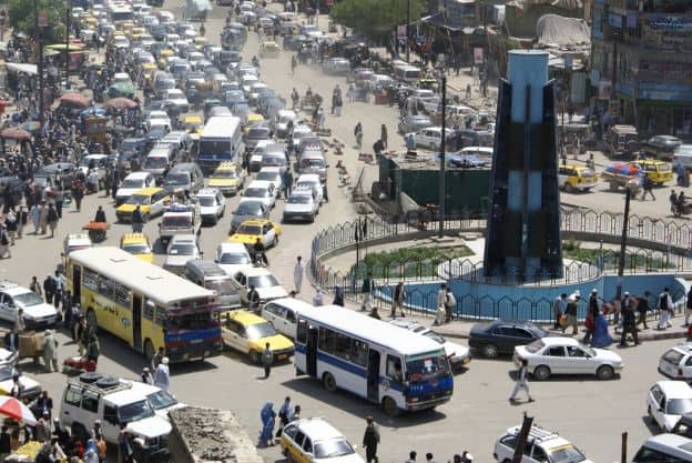Buses, taxis, cars, trucks, and pedestrians move through a busy traffic circle in Kabul, Afghanistan. At the center of the roundabout is an obelisk.