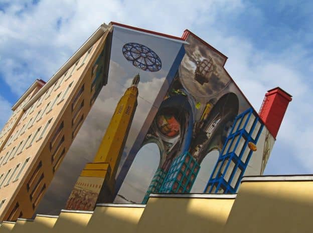A mural in Kotka with a three-dimensional look is painted on the side of a tall building. It shows modern architecture mixed with more traditional styles.