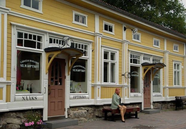 A yellow building with signs in Finnish declares the presence of an optician's shop in Rauma, Finland. Seated on a bench in front of a shop is a modern sculpture of a red-haired woman.
