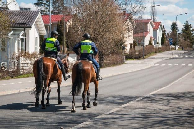 Two Finnish policemen wear reflective vests while patrolling a residential Helsinki neighborhood on horseback. One-storey, red-roofed houses line the street.